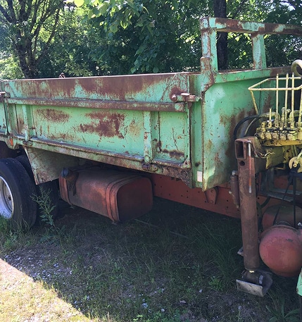 Camion benne vert très rouillé abandonné dans la végétation, réservoir orange, système hydraulique jaune visible.