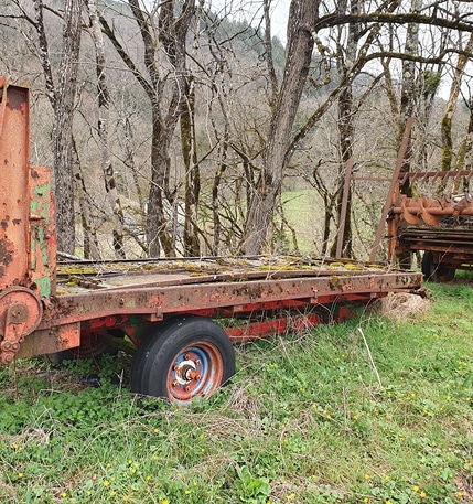 Remorque agricole rouillée abandonnée dans l'herbe, pneu crevé, forêt en arrière-plan.