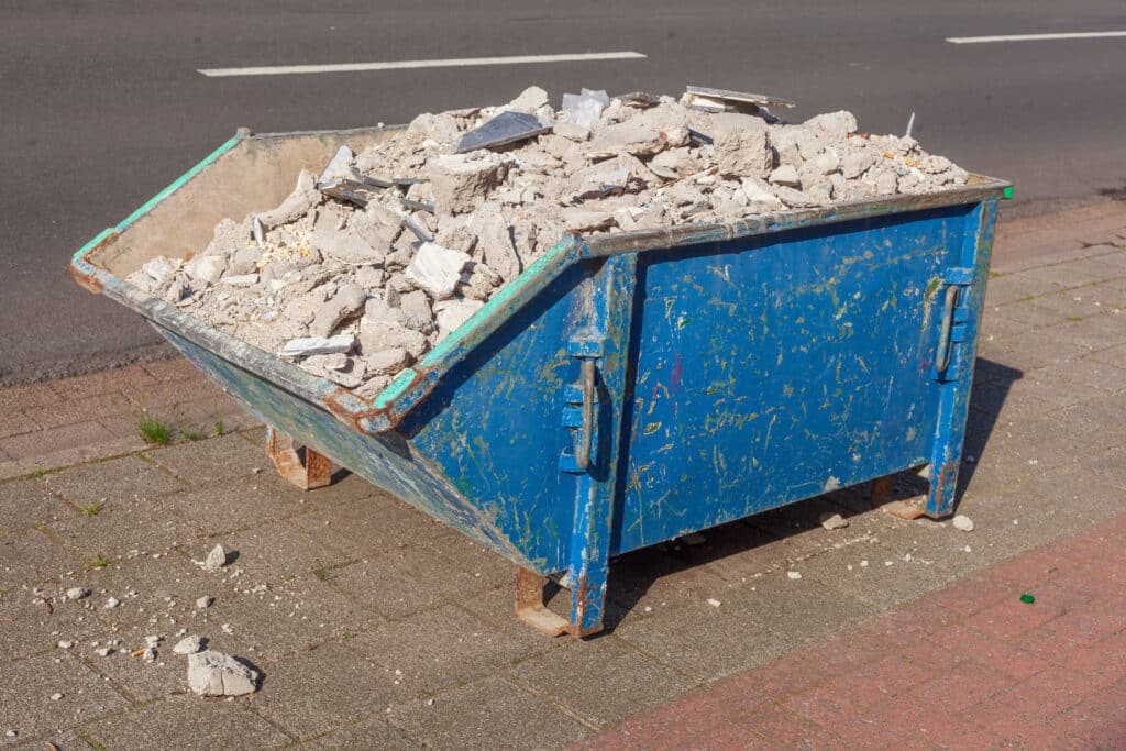 Benne à déchets bleue remplie de gravats, béton et débris de construction, posée sur un trottoir près d'une route.
