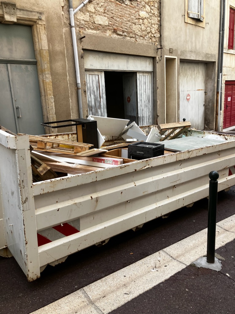 Grande benne blanche pleine de gravats et mobilier usagé sur une rue urbaine, devant des bâtiments anciens. Le numéro 15 est visible.